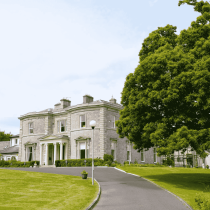 A large, elegant stone mansion with columns at the entrance, surrounded by a well-kept lawn and tall trees, with a curved driveway leading up to the building under a clear sky.